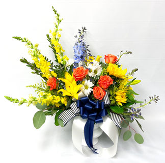 Bright yellow and orange flower arrangement with roses, daisies, blue delphinium accent flowers, eucalyptus and mixed greenery, with a navy blue striped ribbon in a white urn container.