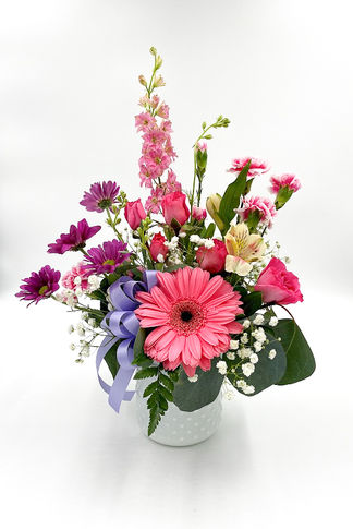 Pink and purple flower arrangement with gerbera daisies, mini carnations, baby’s breath, greenery, and a lavender ribbon in a vase.