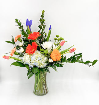 Colorful mixed flower arrangement with orange gerbera daisies, blue iris, peach carnations, white hydrangea, sunflowers, orange tulips, greenery, and assorted accent flowers arranged in an asymmetrical style in a clear glass vase.