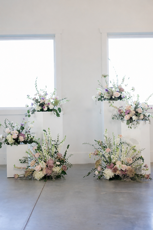 Fresh wedding ceremony flowers arranged on white pedestals in a light, airy setting.