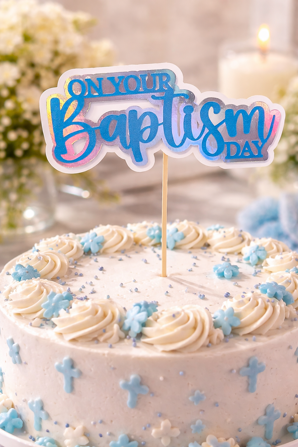 Iridescent blue "On Your Baptism Day" cake topper on a white frosted cake decorated with blue crosses and flowers.