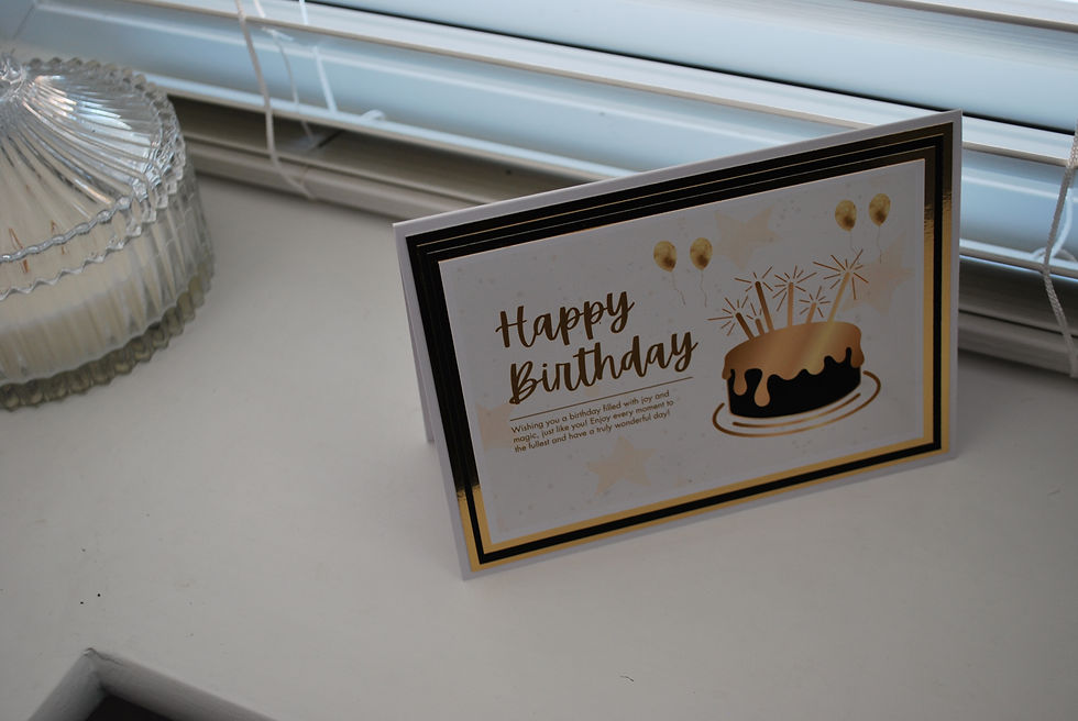 Thumbnail: Close-up of a birthday card with gold foil accents and a cake illustration by a window.