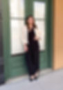 Ashley is smiling and wearing a black jumpsuit with a cream blazer, leaning against a set of green doors.