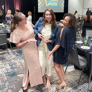 Ashley has an elated expression on her face while holding her Young Professional of the Year award from the Greater Fort Myers Chamber of Commerce award ceremony. She is wearing a navy blue blazer over a white jumpsuit, and posing with two other women.