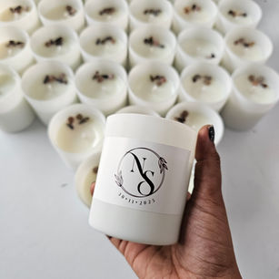 A close-up of a hand holding a clean white frosted glass candle with a round custom botanical monogram label that reads "NS 30.11.2025" against a blurred background of bulk candles.