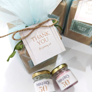 A milestone 50th birthday return gift set featuring a kraft favor box wrapped in soft pastel blue tulle with a classic, minimalist "Thank You for joining us!" tag. Displayed in front are two tiny customized jar candles with gold lids labeled "Nikhil's 50th". One filled with light green wax and the other with pink wax.