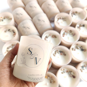 A close-up of a hand holding a personalized frosted glass candle with a custom monogram label, against a background of multiple seashell-topped candles.