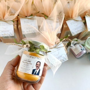 Hand holding a personalized memorial return gift candle with a photo and birth and death dates for Ralph Anthony Smith, rows of wrapped favors in the background