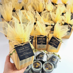 Hand holding a personalized milestone birthday return gift box wrapped in pale yellow tulle with a custom black thank-you tag from Candles with a K.