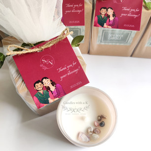 A top-down view of an open frosted glass candle with seashells next to a gift box elegantly wrapped in white net with a magenta wedding favor tag.