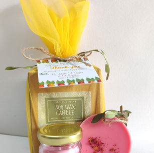 A custom Shashtiabdapoorthi 60th birthday gift set showcasing a jute gift box enclosed in vibrant yellow tulle with a prominent "Soy Wax Candle" label. Arranged neatly in front is a personalized mini jar candle for T.D. Ashok alongside a round pink botanical wax wardrobe sachet.