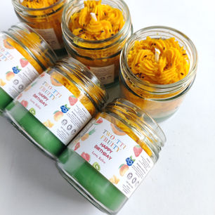 Personalized birthday favor jar candles with vibrant fruit illustrations and custom "Two-tti Fruity Happy Birthday Love, Kriva" labels on layered green and orange soy wax candles.