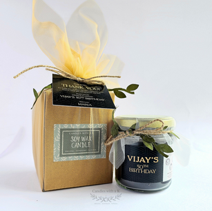 A single personalized "Vijay's 50th Birthday" soy wax jar candle with a rustic leaf accent next to its coordinating cream tulle-wrapped favor gift box against a white background.