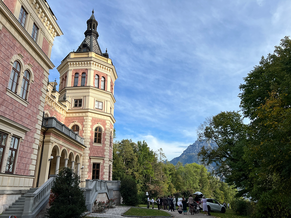Auf der linken Seite das Schloss Traunsee mit Blick auf den Berg Traunstein und den Traunsee. Blauer Himmel, nur ganz leichte Wolkenschleier. Vor dem Schloss steht die Hochzeitsgesellschaft