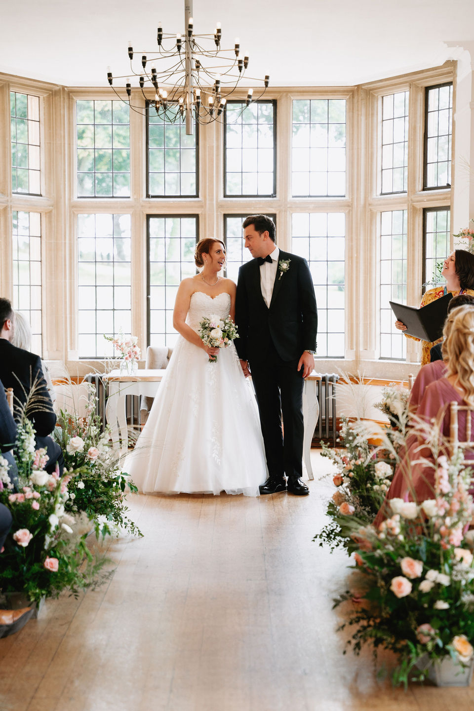 Bride and Groom in front of Registrar table, holding bridal bouquet and wearing buttonhole. Ceremony wedding flowers decorating the aisle. Wedding at Coombe Lodge, Blagdon in Somerset.