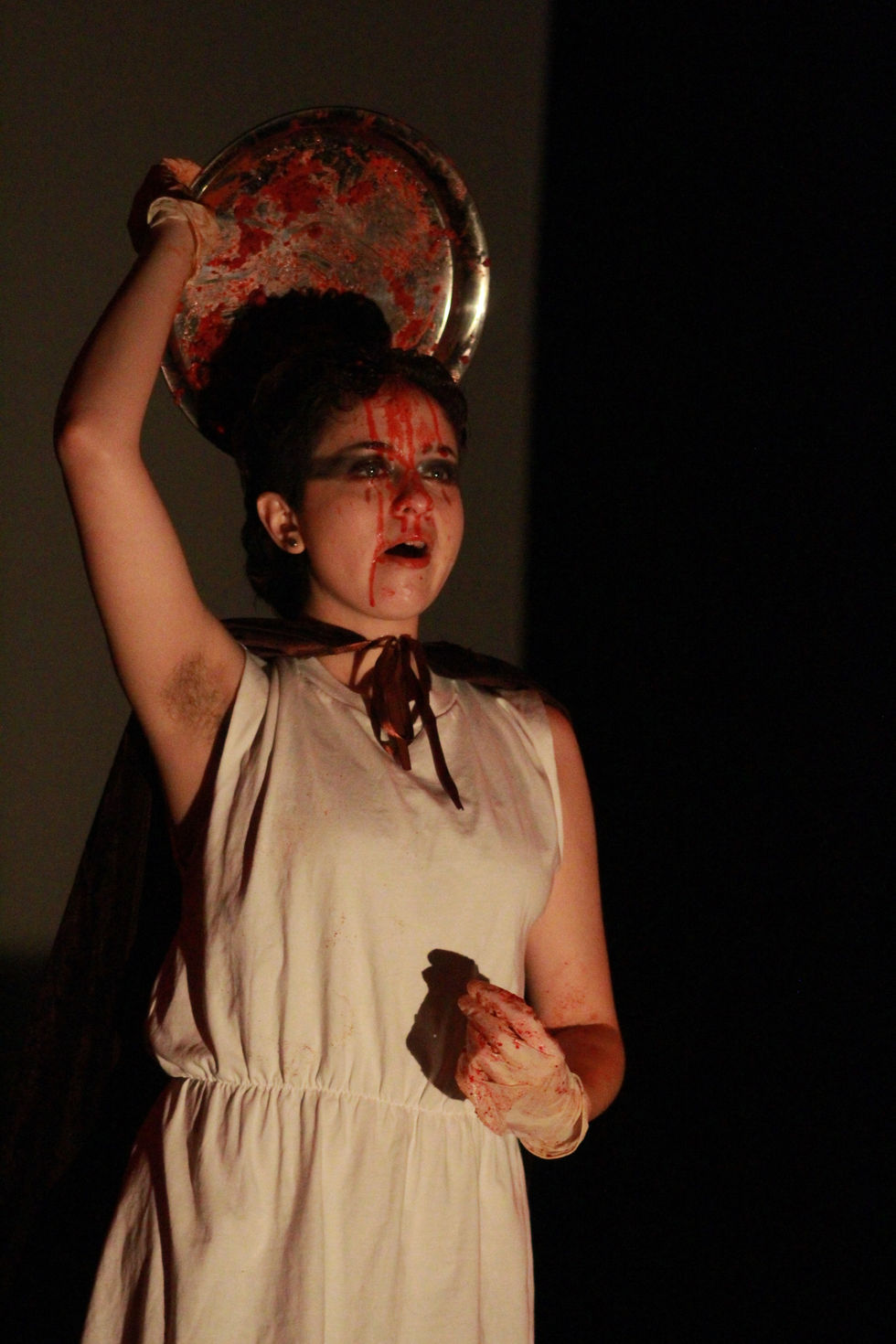 Nadia wears the same costume as described in the first image. Their face and head are covered in fake meat and blood, it drips down their face. They hold the bottom half of the silver dome serving dish behind their head, which is also covered in blood. This positioning creates the imagery of a blood soaked person with a bloody halo.