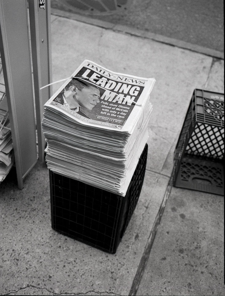 Photographie argentique en noir et blanc, vintage . L'élection de Barack Obama en novembre 2008. La photo représente une pile de journaux posée sur le sol dans une rue de New York,  à l'effigie d'Obama. Le titre : leading man !  