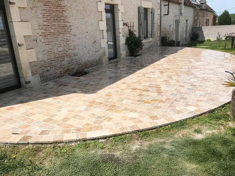 Terrasse pavée incurvée en pierres claires devant une maison rustique avec pelouse.