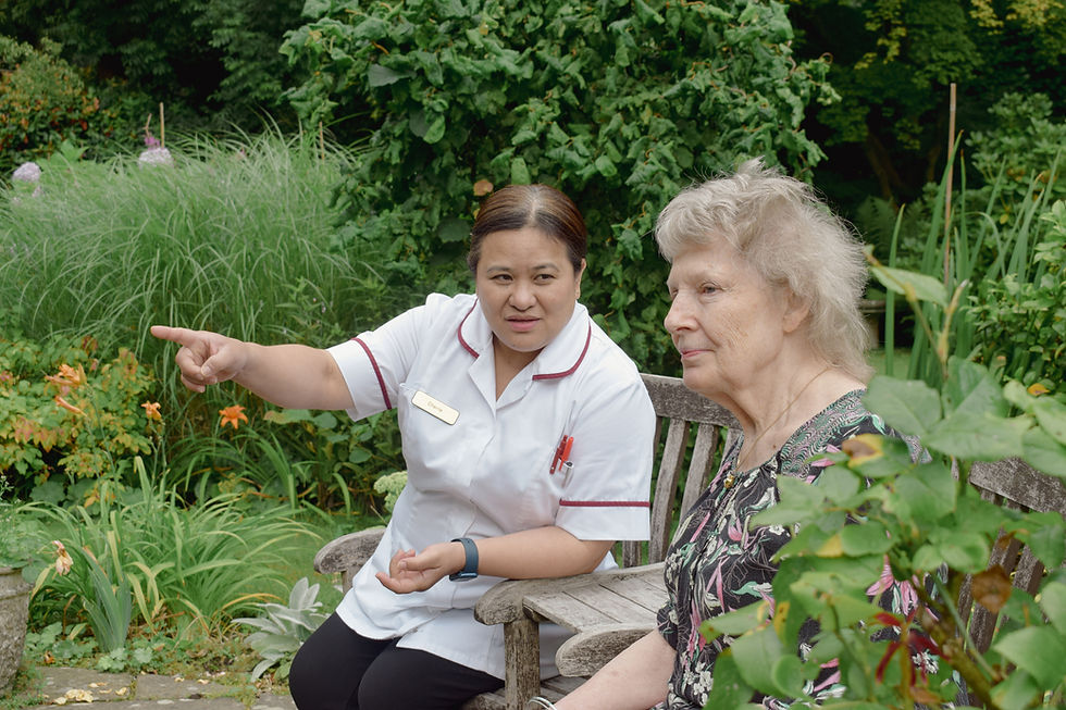 Caregiver pointing while talking to an elderly woman on a garden bench. Lush greenery surrounds them, creating a peaceful atmosphere.