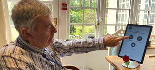 Elderly resident seated by a window, selecting from Loopeli screen displaying options for video calls, photos and radio.