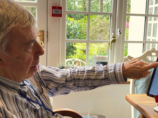 Elderly resident seated by a window, selecting from Loopeli screen displaying options for video calls, photos and radio.