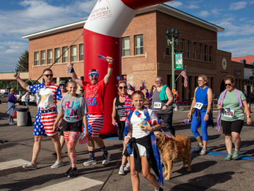 A group of people in patriotic attire participate in a fun run, smiling and holding flags. A red arch and brick building are in the background.