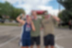 Three people pose outdoors with medals, smiling. One flexes in a USA jersey. Overcast sky, green trees, and a building in the background.