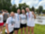 Four smiling girls in white shirts with medals pose outdoors on a grass lawn, displaying excitement. Basketball courts and trees in background.