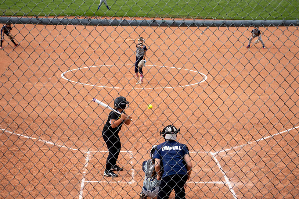 Softball game through fence: pitcher throws ball to batter, umpire watches closely. Players in field, brown dirt, green grass visible.