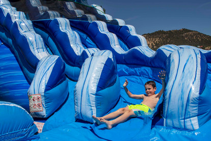 A young boy in neon green swim trunks slides down a large blue inflatable water slide, smiling with arms raised.