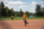 Softball player in yellow catches a ball mid-air on a field. Trees and houses in the background under a partly cloudy sky.