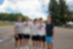 Five young men stand outdoors on pavement, smiling with medals. Trees, cars, and a tent are in the background. Sunny day with clouds.