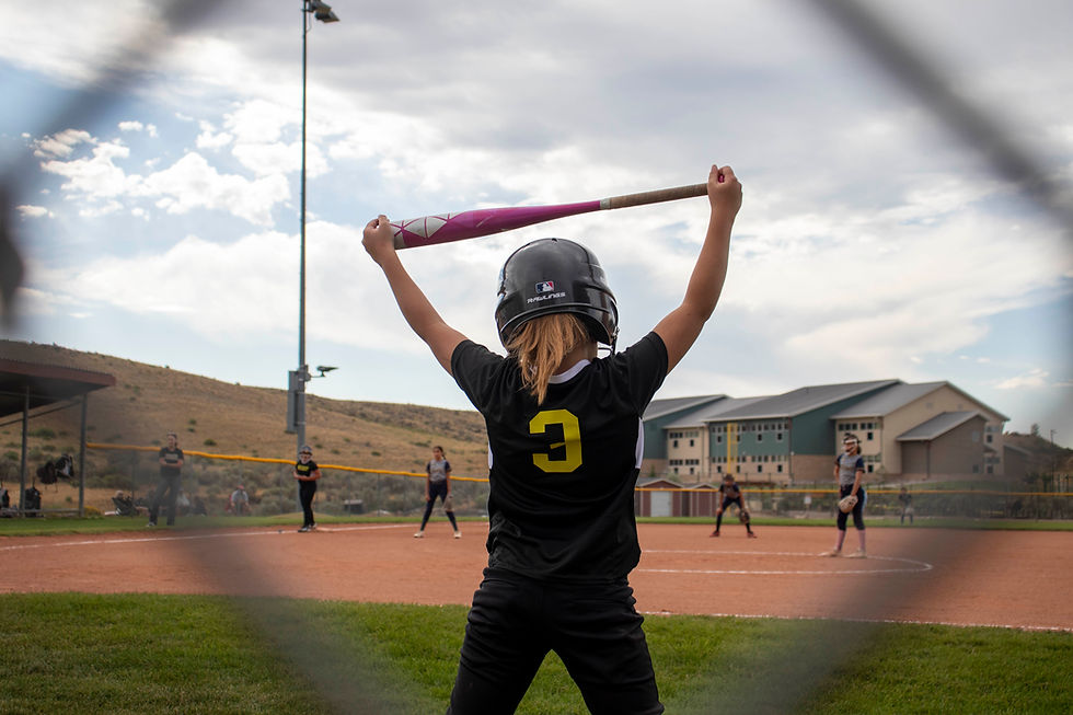 Player in black jersey, #3, holds pink bat ready to swing on a baseball field. Teammates in position, overcast sky, school in background.