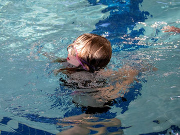 Child wearing goggles swims in a pool, partially submerged. Blue water reflects light. Another hand visible reaching in from the right.