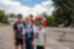 Three boys with medals pose on a basketball court, smiling. Background: trees, building, red arch with "ERBM." Cloudy sky.
