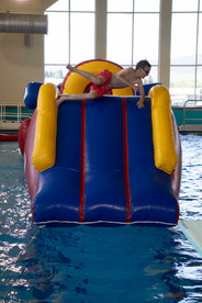 Boy in red swim trunks and goggles climbs across the top of a blue and yellow inflatable structure floating in an indoor pool.