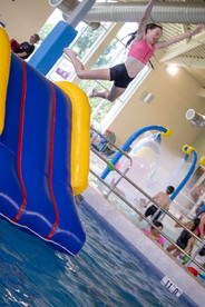 Young girl in a pink top and black swim shorts mid-jump from a large inflatable slide into an indoor pool, arms extended and smiling.