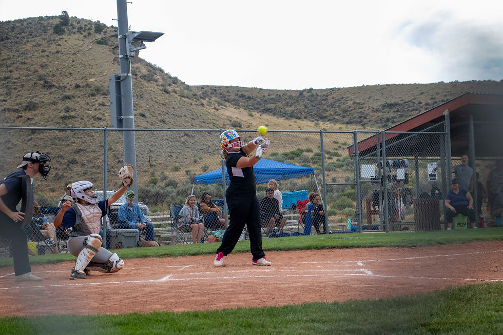 Child in colorful helmet swings bat at softball on field, with catcher ready. Hilly background; spectators watch from behind a fence.