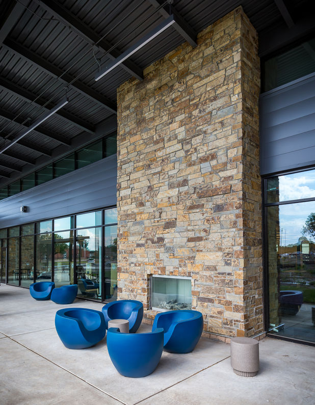 Outdoor lounge area at Newton Public Library featuring a stone fireplace, modern blue sculptural chairs, and a covered patio. Large glass windows provide a seamless connection between indoor and outdoor spaces.