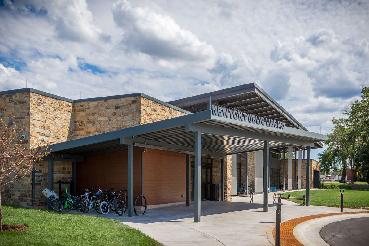 Main entrance of Newton Public Library featuring a covered walkway, stone and metal façade, bicycle racks, and outdoor seating. The modern architectural design integrates natural materials with contemporary elements.