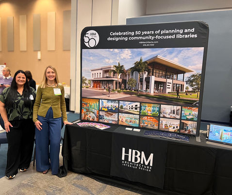 Principal and Interior Designer Brooke Breiner and Interior Designer Mackenzie Marinelli at NEFLIN in Jacksonville, Florida, April 2026. Pictured at their booth, which displays a large backdrop featuring an array of HBM Architects' Florida Library projects.