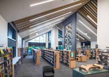 Looking through the main collection area at Clarksville-Montgomery County Public Library North Branch, designed by HBM Architects. Beneath high sloped ceilings with acoustic panels, rows of book stacks with warm wood and colorful resin end panels are surrounded by varied seating options, including window counters and quiet lounge nooks. A geometric acoustic divider screen in shades of blue, tan, and white provides see-through sound absorption between zones.