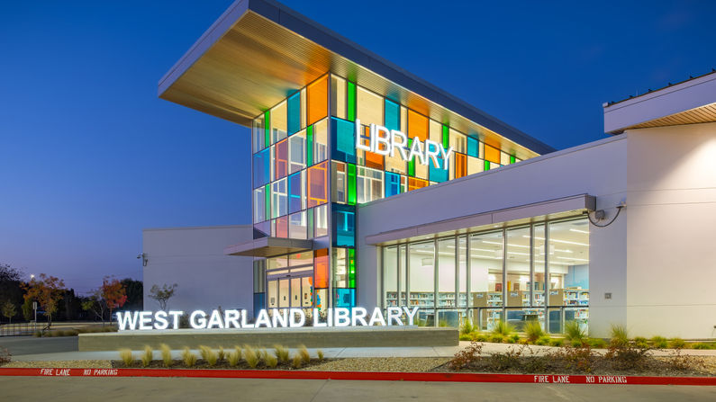 Exterior night view of the new West Garland Library in Garland, Texas. The entry features a central volume of colored glass that reflects onto the floor and walls of the space like a kaleidoscope. These blues, greens, and coral colors are further used throughout the interior to guide patrons to the adult area, teen and children's area, or large meeting room from this main spine. At night, these coloful glass windows are higlighted against the dark sky, adding a vibrant glow to the night sky.