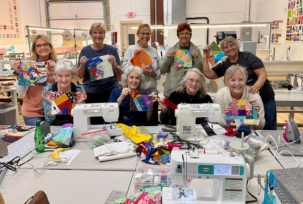 Nine women in a sewing studio holding colorful quilt pieces, smiling. Sewing machines and vibrant fabric on tables. Bright, creative atmosphere.