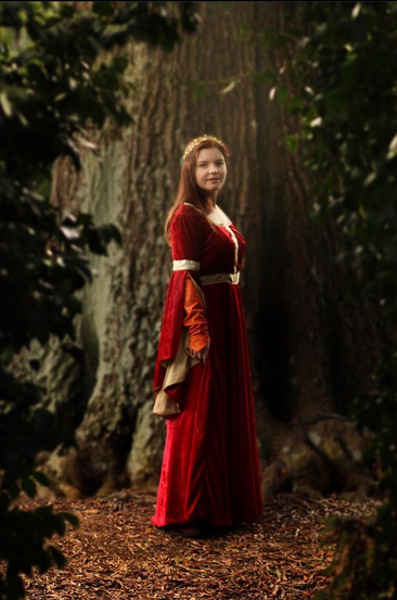 person in a red medieval-looking dress with gold accents and a crown standing in front of a tree. bushes frame the photo