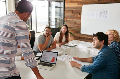 Colleagues engaged in discussion during a team meeting, gathered around a conference table with a laptop displaying a presentation graph.