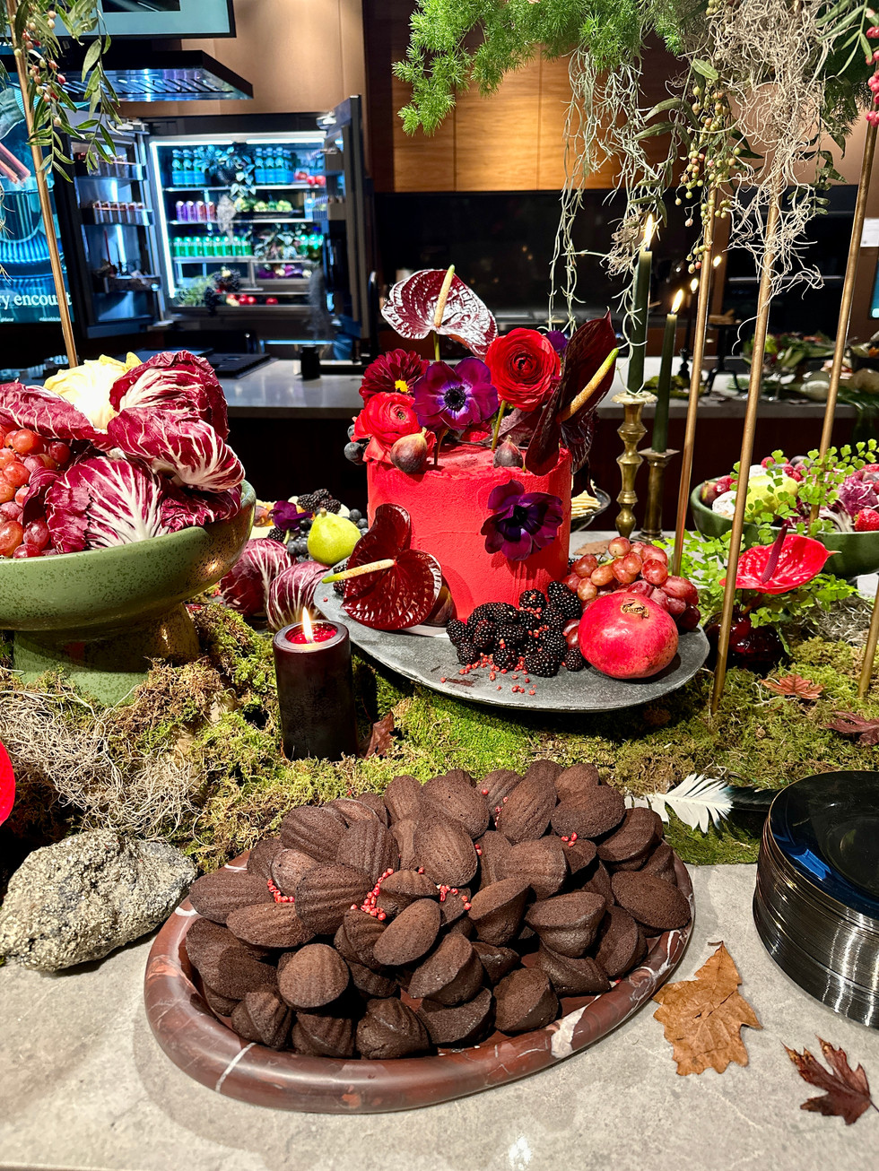 A vibrant tablescape featuring dark chocolate madeleines and a striking red floral arrangement with pomegranates, grapes, and candles, creating a rich, dramatic setting.
