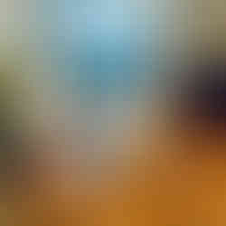 A bright room with wooden floors, glass doors leading to a balcony, potted plants, and minimal furniture including a small white ottoman and a black dresser.