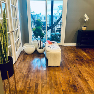 A bright room with wooden floors, glass doors leading to a balcony, potted plants, and minimal furniture including a small white ottoman and a black dresser.
