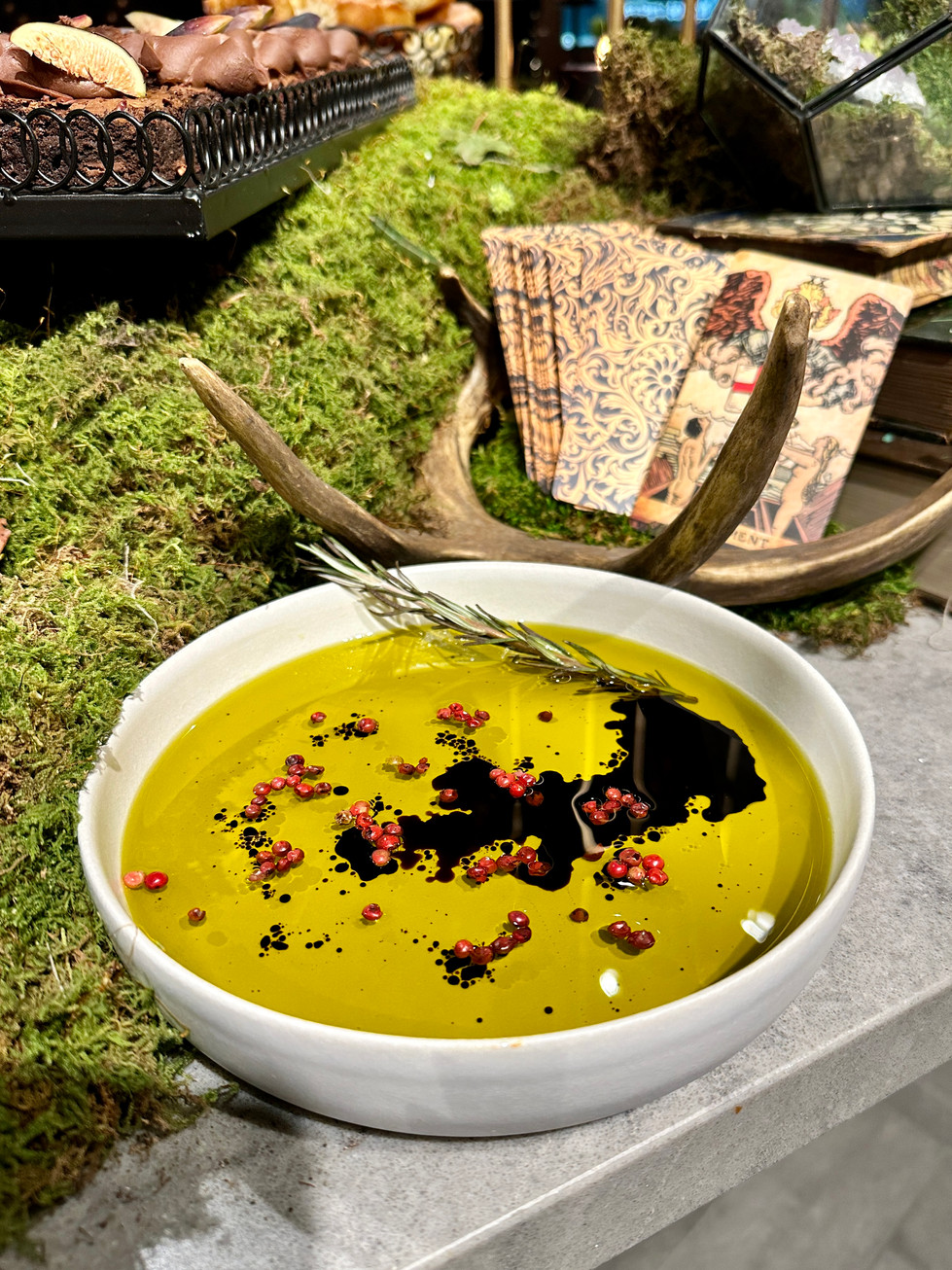 A close-up of a dish filled with olive oil and balsamic vinegar, garnished with rosemary and pink peppercorns, set on a moss-covered table with rustic décor.